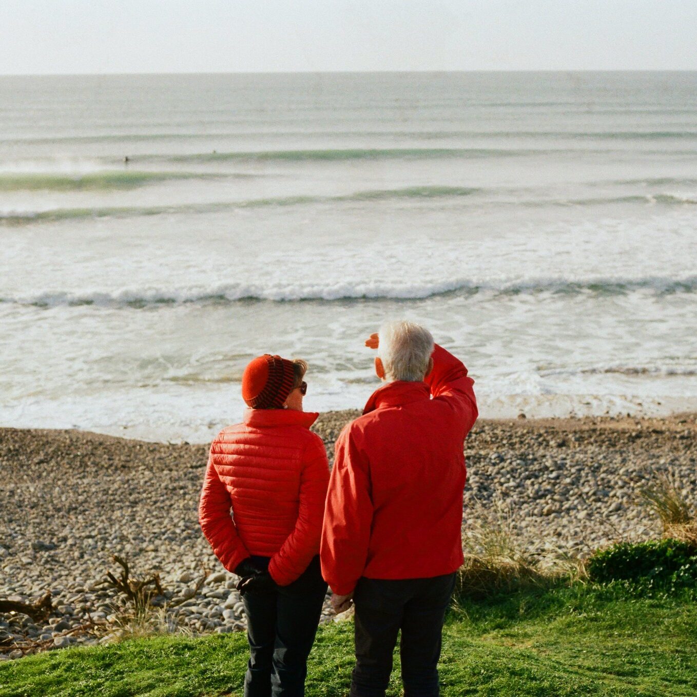 Un couple de personnes âgées regarde au loin la mer et l'horizon depuis une plage. Ils portent tous les deux un manteau rouge. La photo dégage une atmosphère positive et optimiste. 